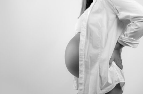 Schwangere Frau mit geöffneter weißer Bluse hält sich die Hände im Rücken